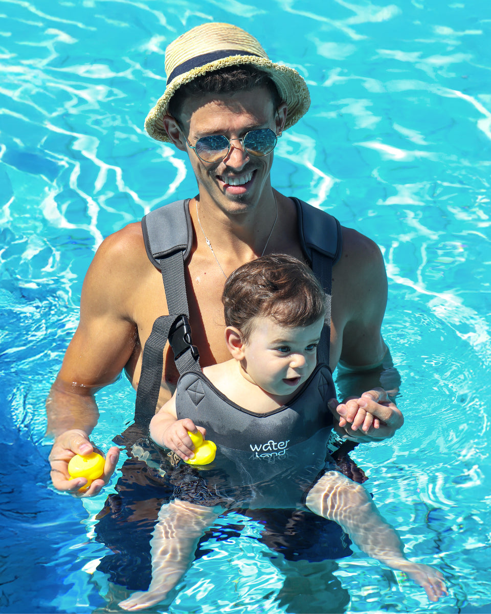 A parent standing waist-deep in a bright blue pool, a baby in an Urban Grey WaterLand carrier and a yellow rubber duck