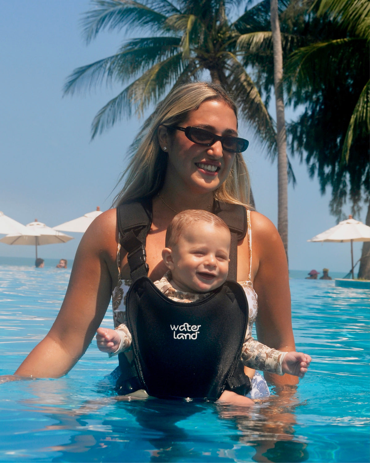 A parent standing waist-deep in a hotel pool under palm trees, wearing a laughing baby in a Jet Black WaterLand carrier
