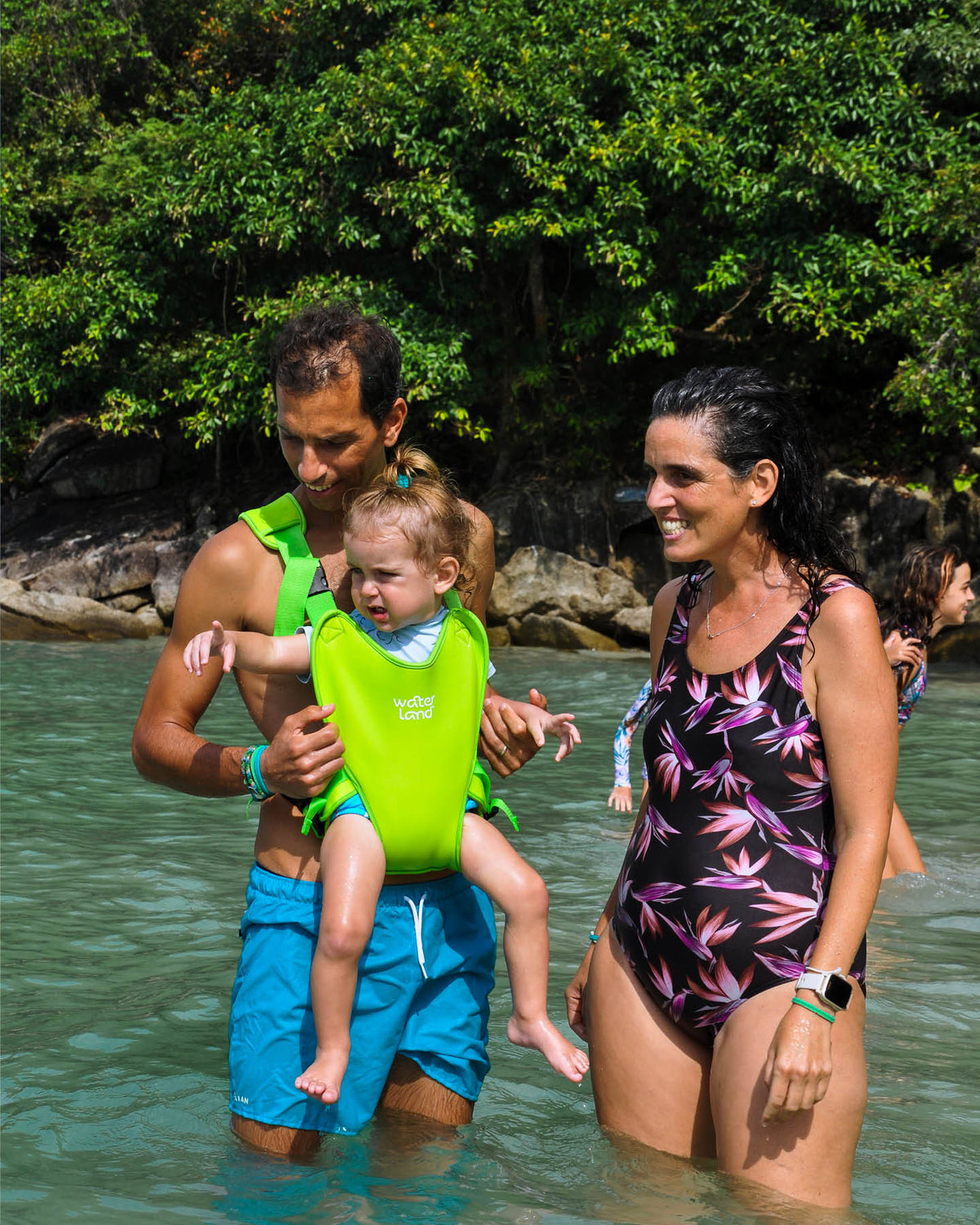 A father and mother wading hip-deep in a turquoise sea, their baby in a Keylime Green WaterLand carrier on his chest