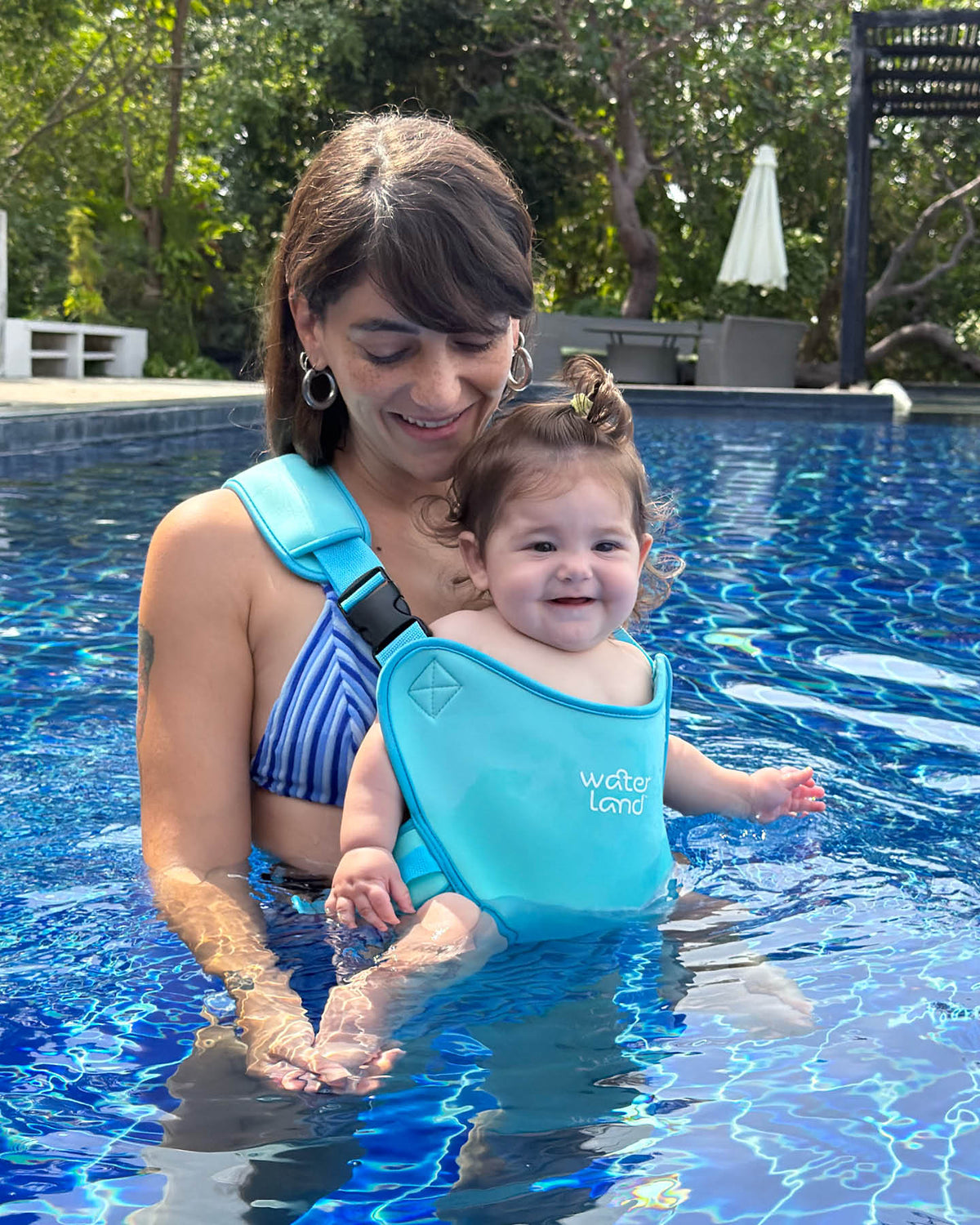 A mother stands waist-deep in a blue pool, her smiling baby close in a Light Blue Sky WaterLand carrier