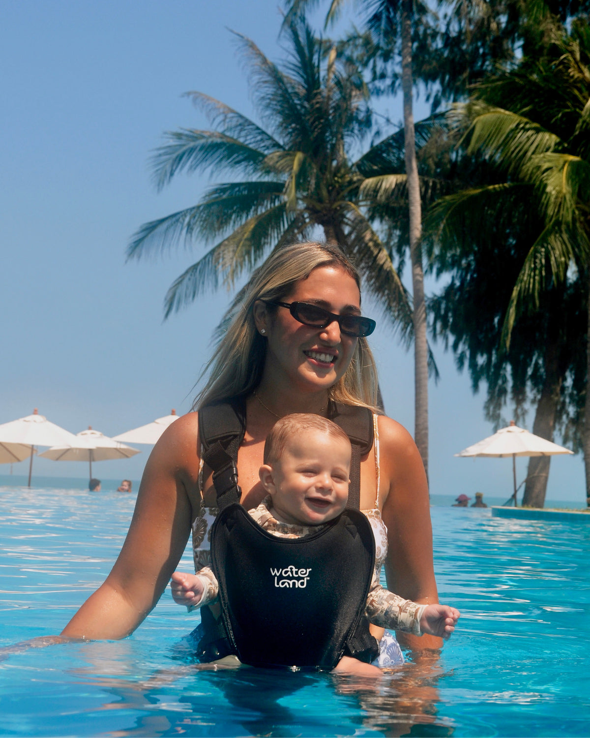 A parent standing waist-deep in an infinity pool under palms, a baby in a Jet Black WaterLand carrier
