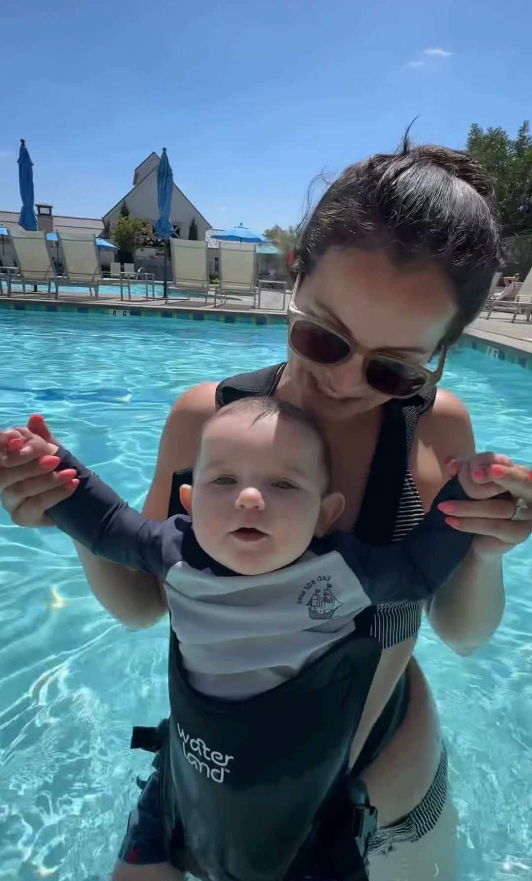 Smiling woman in a pool holds both hands of a baby in a Jet Black carrier, who looks at the camera.