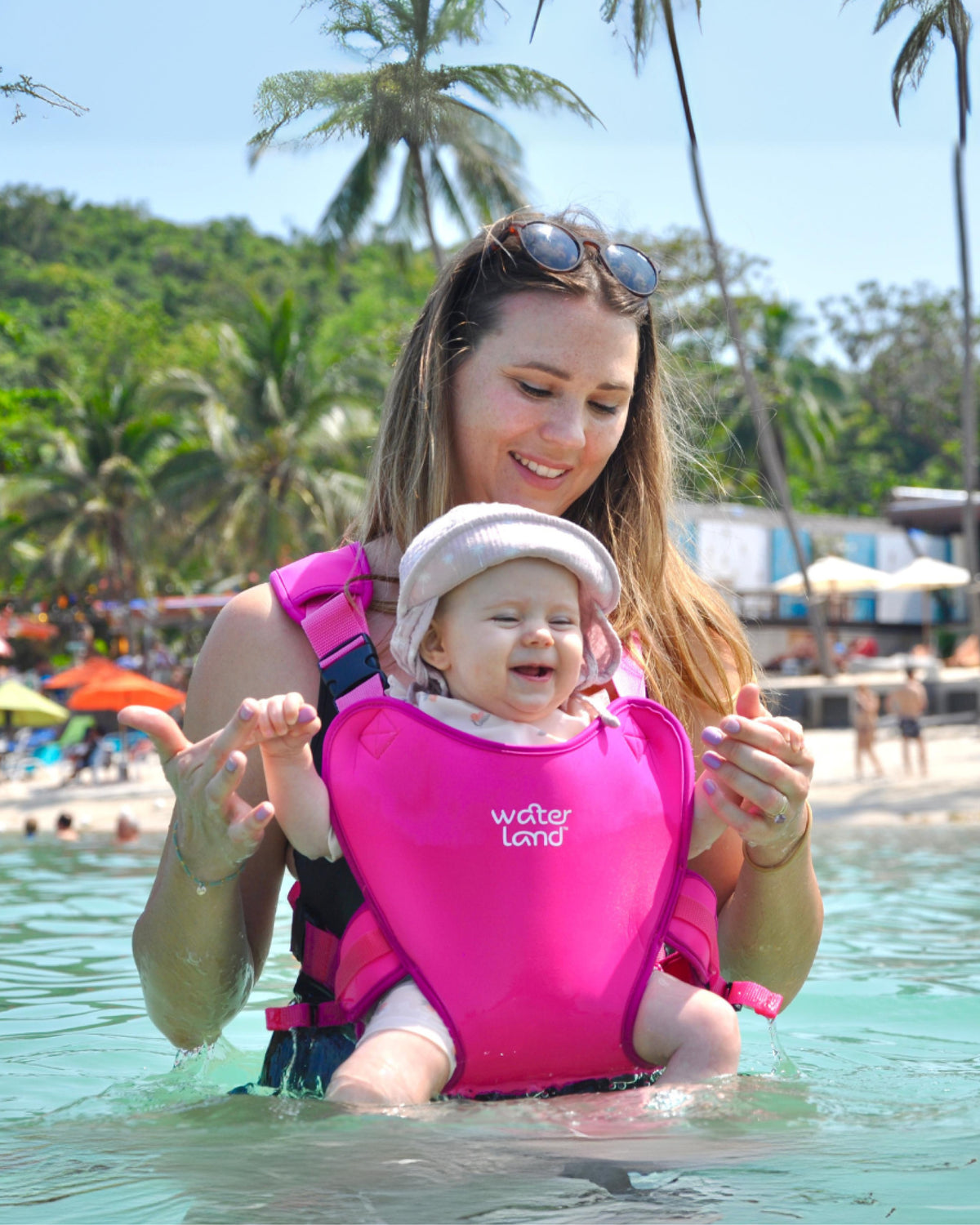 A parent standing in calm, shallow sea water, wearing a smiling baby in a Super Pink carrier