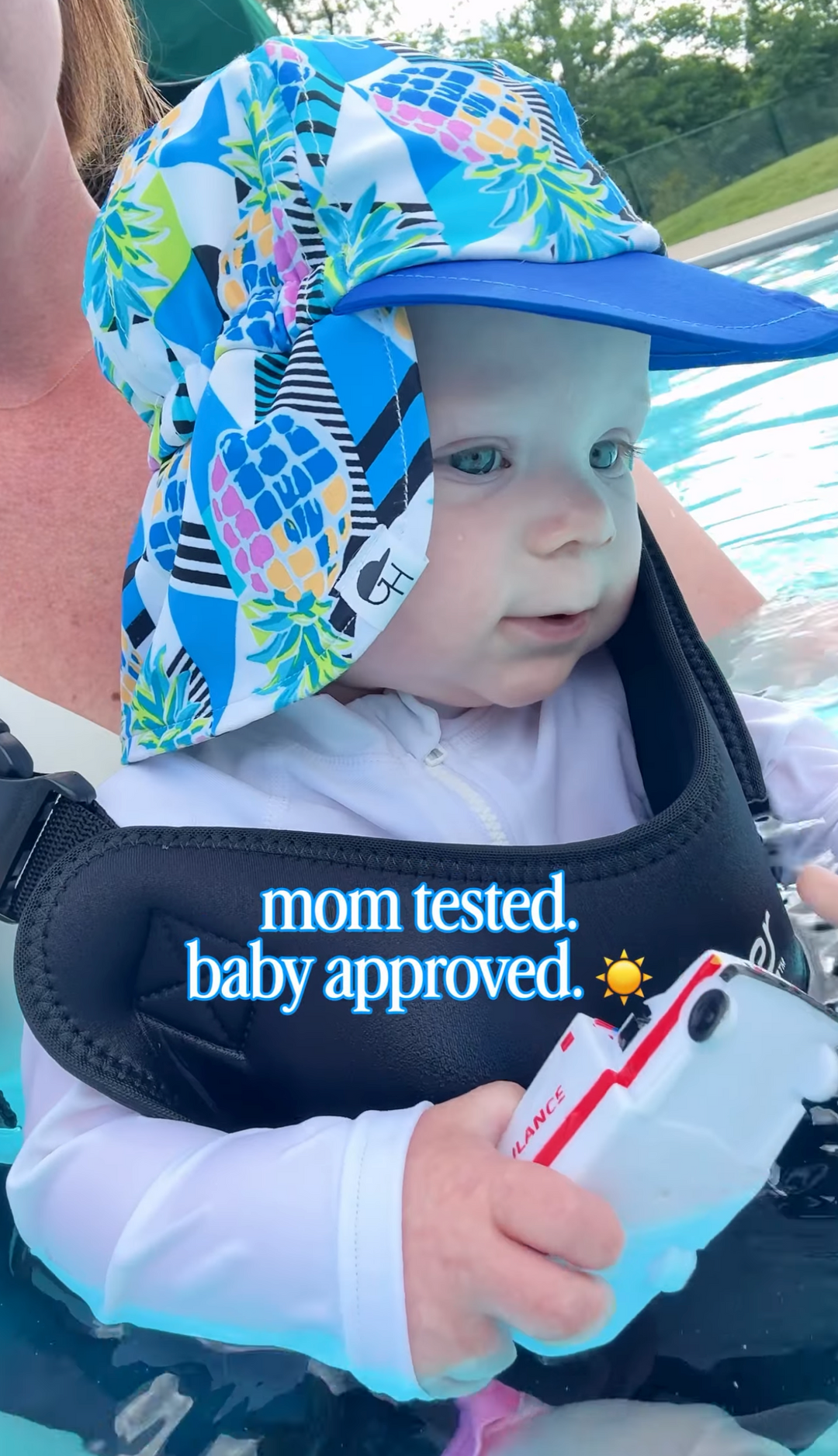 Baby in a Jet Black carrier in a pool, wearing a pineapple-print sun hat and holding a toy ambulance. Text on photo: “mom tested. baby approved.”