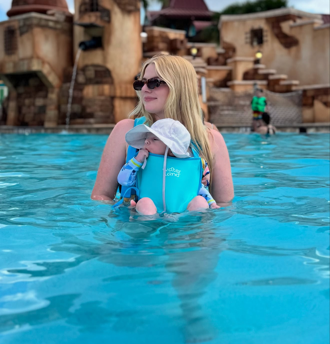 Woman in sunglasses stands waist-deep in a pool in a Light Blue Sky carrier, baby in a white sun hat facing outward.