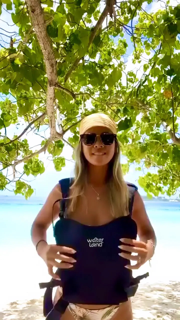 A parent in a sun hat on a dry beach wearing a navy WaterLand carrier, steadying it with both hands